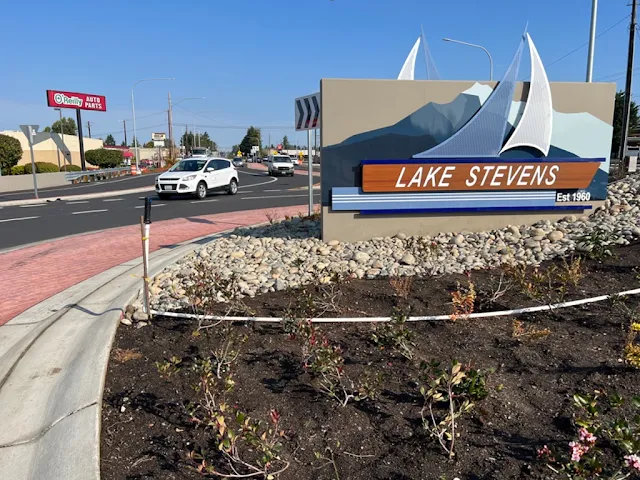 Vehicles enter a roundabout in a business area of Lake Stevens along State Route 9. A sign that reads “Lake Sevens” with a sailboat and landscaping are in the center of the roundabout.