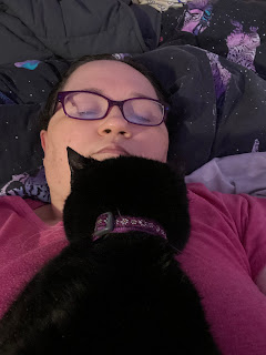 A selfie of a middle aged, brunette, bespectacled, white woman, wearing a pink T-shirt and laying down on her back in bed; using a bunched up black duvet as a pillow. A black cat wearing a pink collar is laying on her front on the woman's chest - between the woman's boobs - with the top of her head resting on the woman's chin. The woman's eyes are open just a crack and looking at the camera, and she has a neutral facial expression.