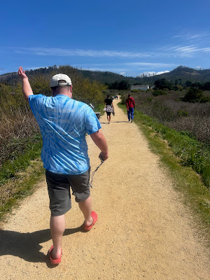 Photo of Leo from behind on a trail to the beach. He is wearing a blue tie-dyed shirt with yellow text that reads, "Bench hiking is beautiful. Catch up if you want". Photo of Leo from behind on a trail to the beach. He is wearing a blue tie-dyed shirt with yellow text that reads, "Bench hiking is beautiful. Catch up if you want"..