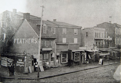 a black and white photo of a dirt street with a set of trolley tracks in it lined with ramshackle looking buildings. circa 1870?