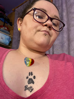 A selfie taken by a brunette, bespectacled, middle aged white woman wearing a pink v-neck T-shirt. She's wearing a necklace with a large pendant; the pendant is heart-shaped clear acrylic, in the background of the pendant is a rainbow with vertical stripes, and in the foreground on the left are some grey speckles, in the middle is a dried forget-me-not (a small blue flower), and on the right is a lock of black fur. She has a black tattoo in the middle of her chest, over her heart, of a cat's paw print, and the name 'Betty' beneath.