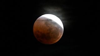 A photo of a full moon captured during a lunar eclipse in a black sky. Earth's shadow covers the moon, turning it a dull crimson, while a thin crescent is visible to the upper right of the lunar disk.