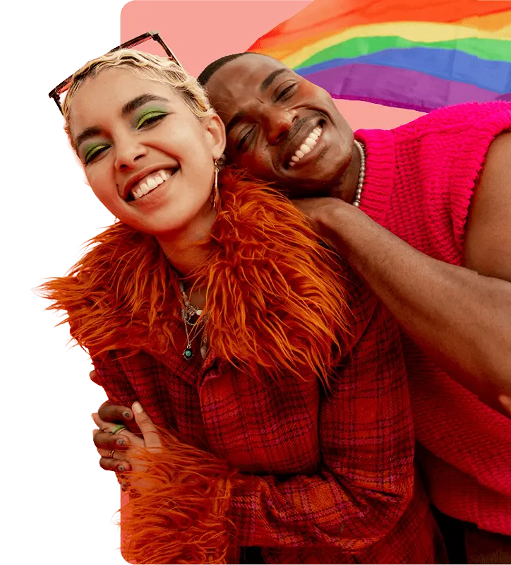 A man and woman hug happily, standing in front of a bright rainbow flag that represents LGBTQ+ pride.