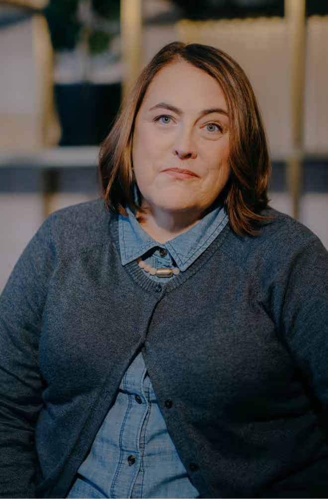 Portrait of a woman with shoulder-length brown hair wearing a blue denim shirt and dark gray cardigan, sitting indoors with shelves and plants in the background.