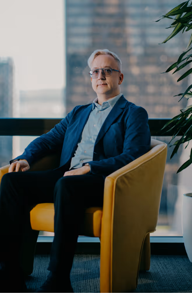 Man with light hair and glasses wearing a blue blazer sitting in a yellow armchair by a large window overlooking city buildings.
