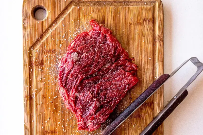 An image of a flank steak on a brown cutting board with a set of tongs next to it.