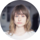 Portrait of a young woman with shoulder-length light brown hair and bangs, wearing a white top and earrings, against a blurred indoor background.