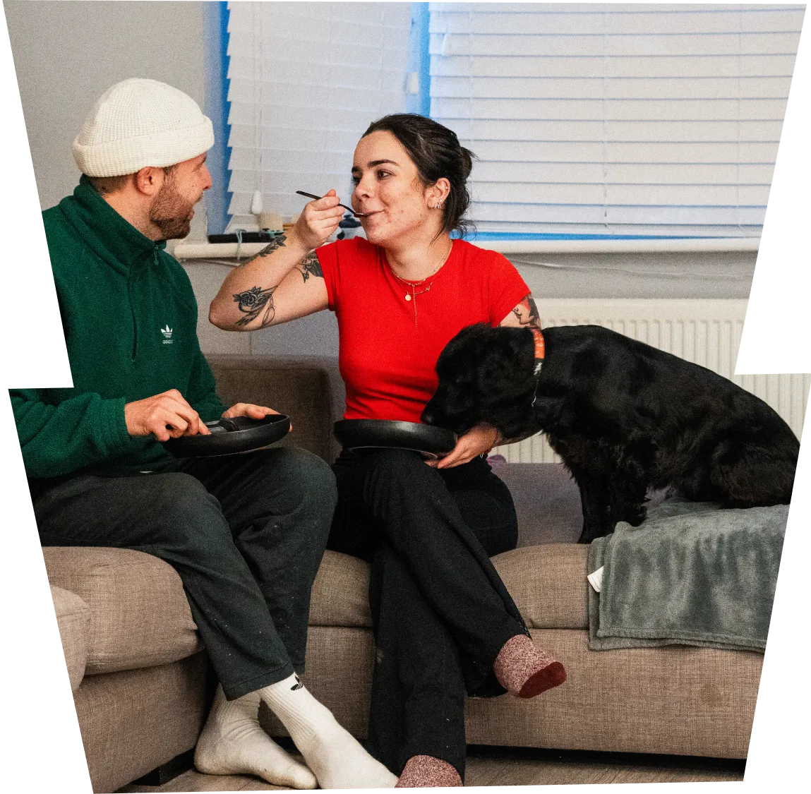 A man in a green hoodie and white beanie and a woman in a red shirt sitting on a couch eating, with a black dog beside them.