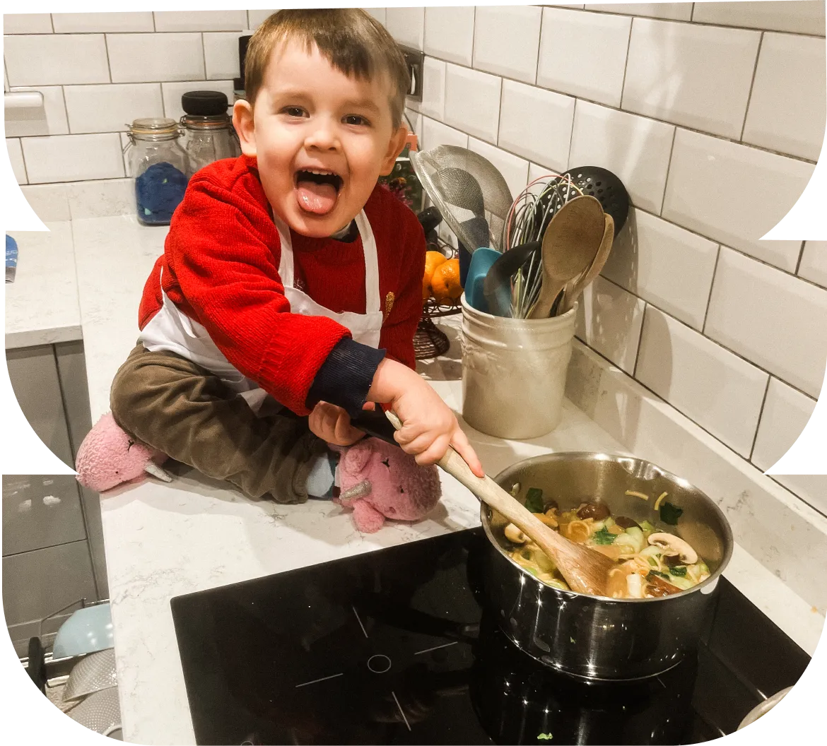 Young child in a red sweater and white apron stirring a pot of soup with mushrooms on a stove, sticking out their tongue playfully.
