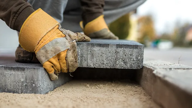 Person wearing gloves placing a gray paving stone onto a sand base during outdoor pavement installation.