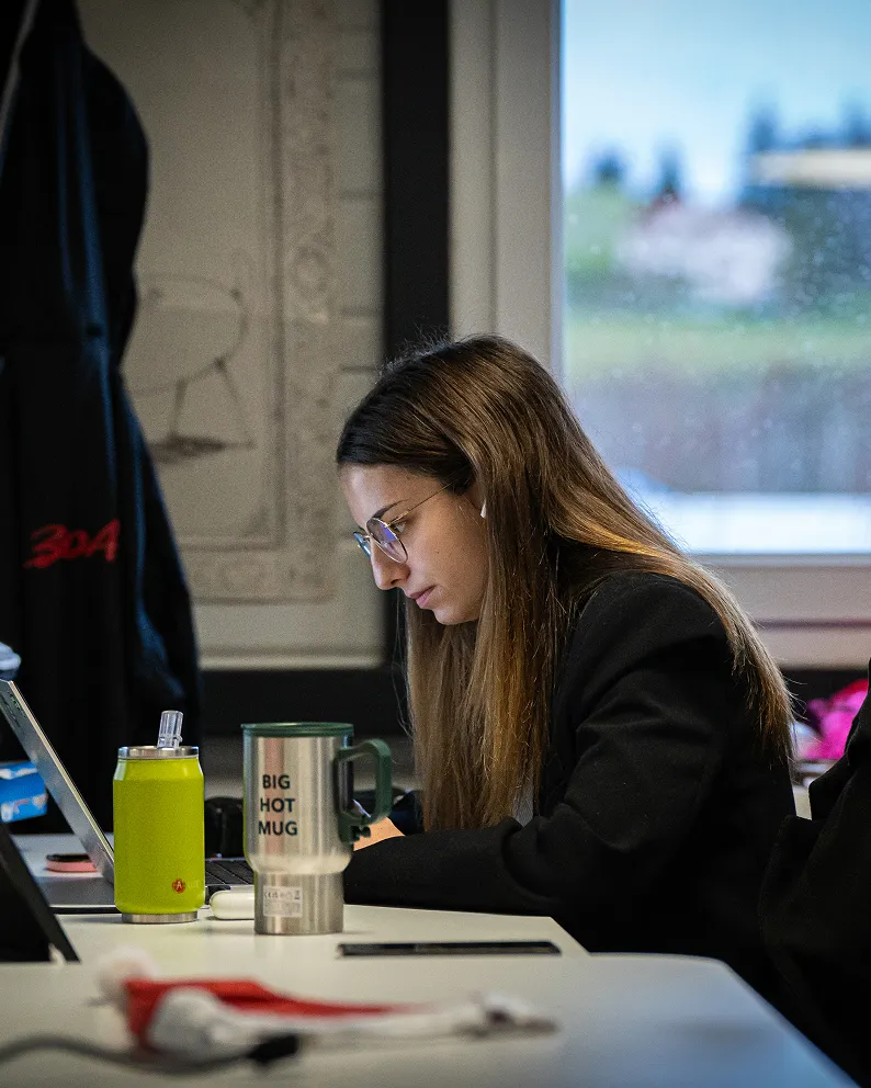 Jeune femme à lunettes travaillant avec concentration sur son ordinateur portable dans un bureau, avec un mug isotherme au premier plan