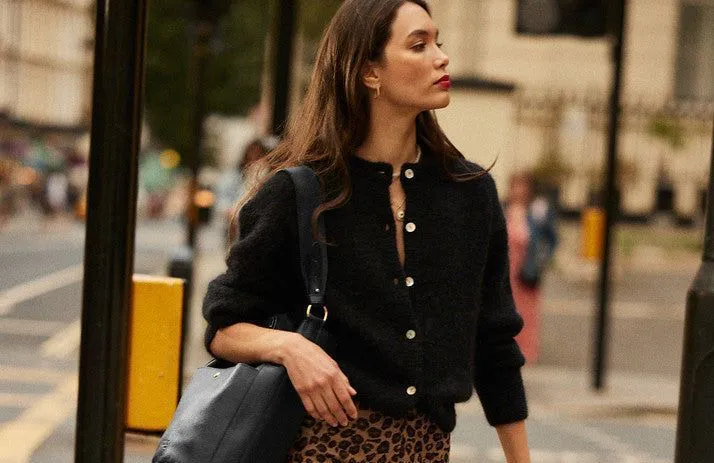 Femme portant un pull noir avec boutons et une jupe léopard, marchant en ville avec un sac à main noir.
