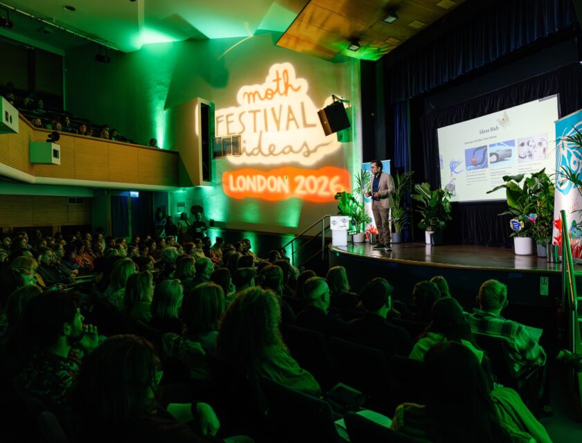 MOTH Festival of Ideas, London 2026 César Rodríguez-Garavito presents onstage to a full audience during the MOTH Festival of Ideas London 2026. Green lighting fills the auditorium while a large illuminated festival projection appears on the wall beside the stage. Presentation slides are displayed on a screen surrounded by plants and festival banners.