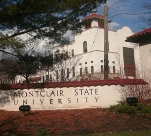 White stucco entrance and tower at Montclair State University, with the university name displayed on a low wall in the foreground and trees surrounding the building.