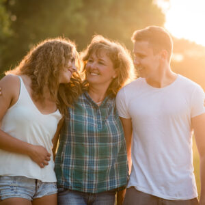 Photograph of a teen girl and boy walking with an adult woman and hugging. The sun is behind them and they are outdoors in an area with trees.