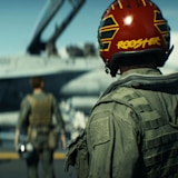 Rear view of a fighter pilot wearing a red flight helmet and flight suit on an aircraft carrier deck, facing parked fighter jets while flight crew members walk in the background.