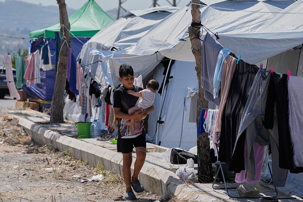 A young boy walks along a dirt pathway next to a concrete curb, carefully cradling a baby in his arms. They are in a makeshift camp lined with rows of white and green canvas tents pitched on a paved area. Clotheslines strung between young trees and tent frames are filled with hanging laundry, including towels and garments, drying in the sun.