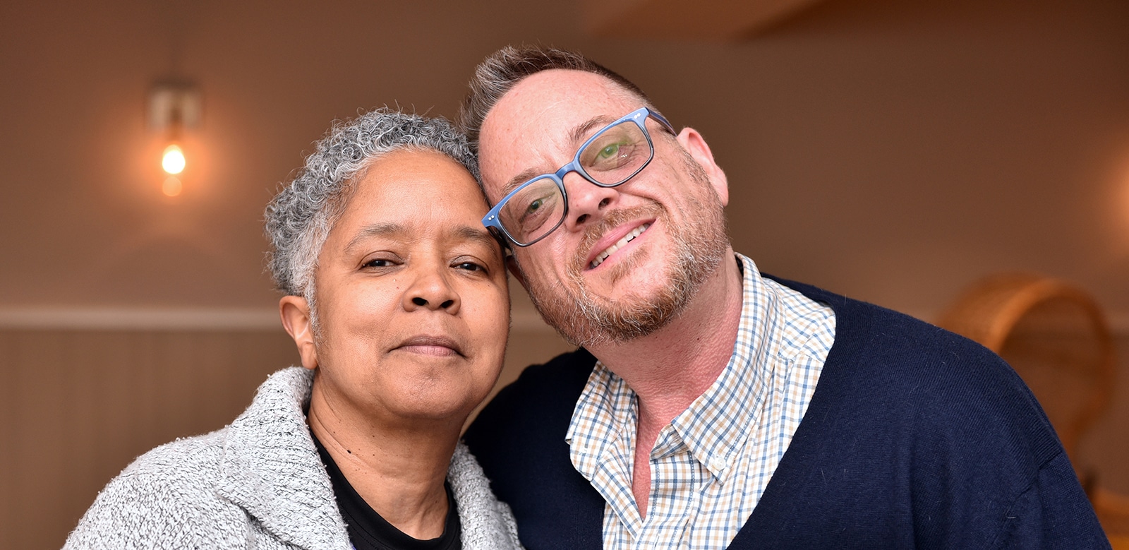 Eva, an older Black woman with grey curly short hair, and George Henry, a white trans man with glasses lean into each other and smile comfortably