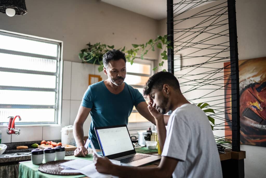 a person looking pensive seated at a computer, their partner looking at them seriously
