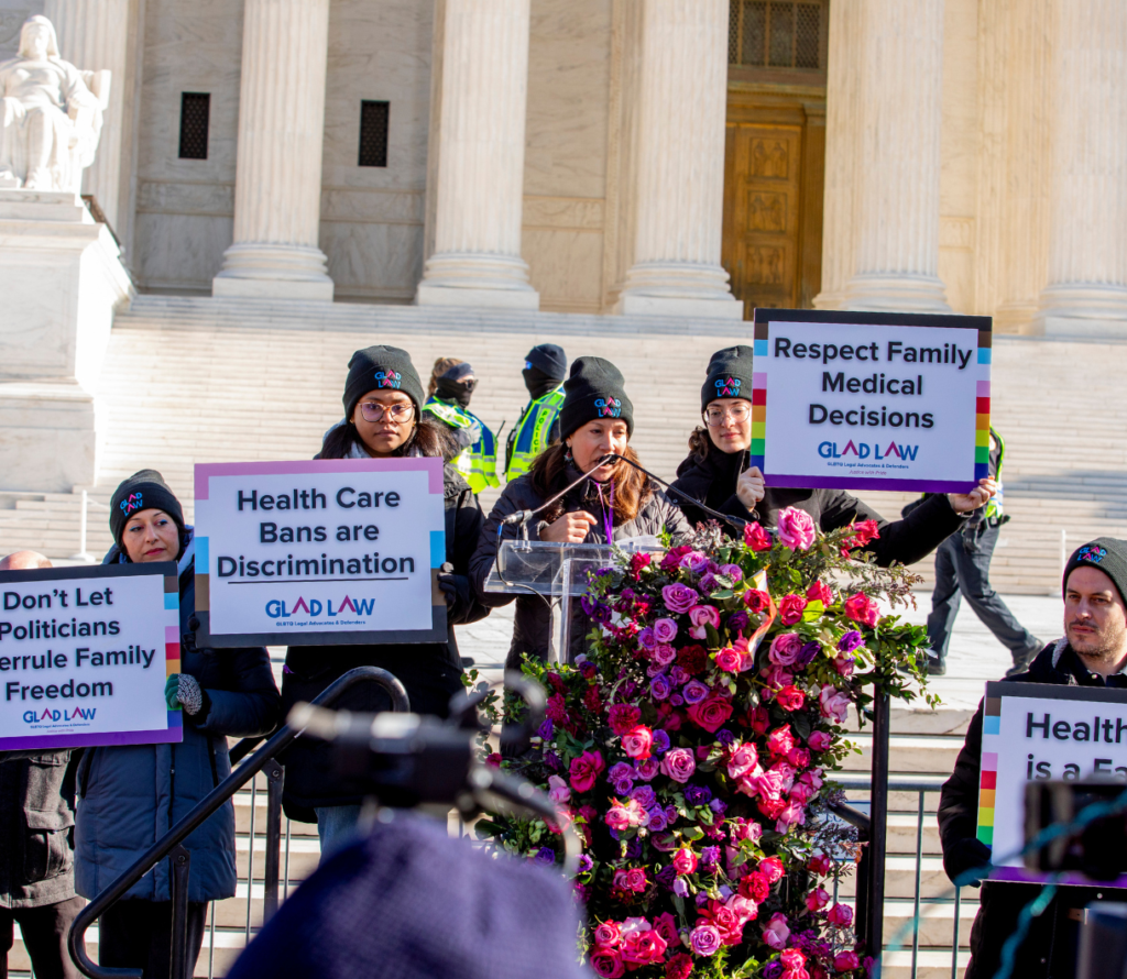 GLAD Law attorney Lisa Rodriguez-Ross speaking at the podium in front of the Supreme Court, surrounded by GLAD Law staff holding signs supporting health care for transgender adolescents.
