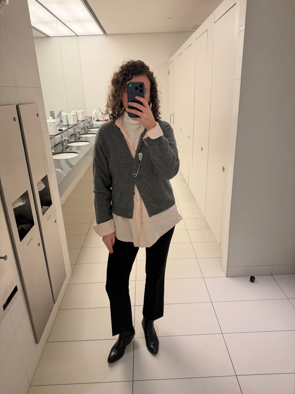 Person taking mirror selfie in a restroom while wearing layered clothing. Person taking mirror selfie in a restroom while wearing layered clothing.