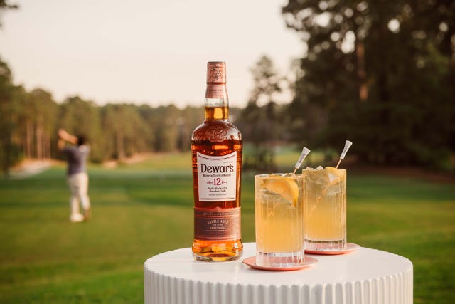 A bottle of Dewar's 12-Year-Old Scotch whisky beside two cocktails on a table with a golfer in the background. A bottle of Dewar's 12-Year-Old Scotch whisky beside two cocktails on a table with a golfer in the background.