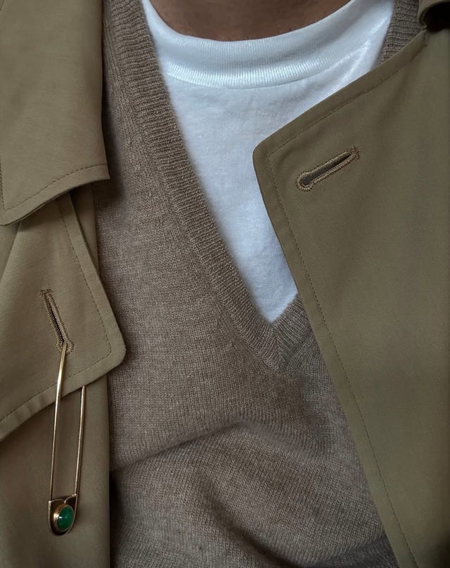 Layered outfit featuring a white shirt, beige sweater, and tan coat with gold safety pin detail. Layered outfit featuring a white shirt, beige sweater, and tan coat with gold safety pin detail.