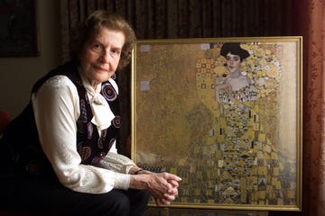 An elderly woman sitting next to a framed portrait in a well-decorated room. An elderly woman sitting next to a framed portrait in a well-decorated room.
