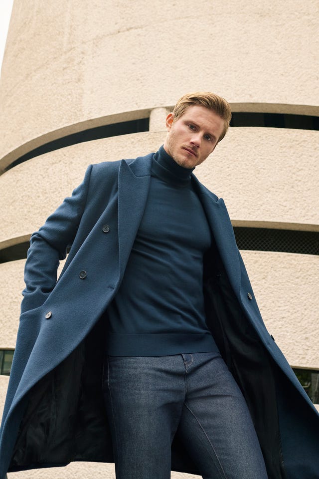 Man in a blue coat and turtleneck standing in front of a textured concrete wall. Man in a blue coat and turtleneck standing in front of a textured concrete wall.
