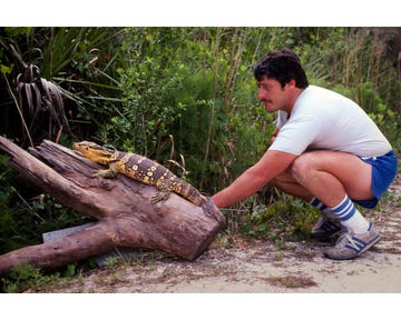 Tom Crutchfield 'Monsters of God' man crouching beside a large, colorful lizard on a log in a natural outdoor setting. Tom Crutchfield 'Monsters of God' man crouching beside a large, colorful lizard on a log in a natural outdoor setting.