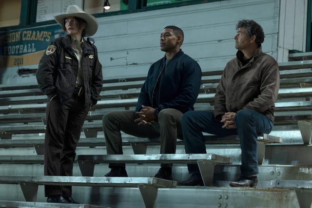 Three individuals seated on bleachers, with one person in a sheriff's uniform wearing a cowboy hat. Three individuals seated on bleachers, with one person in a sheriff's uniform wearing a cowboy hat.