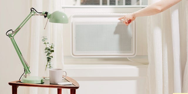 A person adjusting a white air conditioner beside a green desk lamp and a mug on a wooden table by a window. A person adjusting a white air conditioner beside a green desk lamp and a mug on a wooden table by a window.