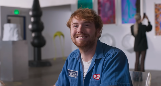 Smiling person with reddish-brown hair wearing a blue shirt, sitting in an art gallery with colorful artworks in the background. Smiling person with reddish-brown hair wearing a blue shirt, sitting in an art gallery with colorful artworks in the background.