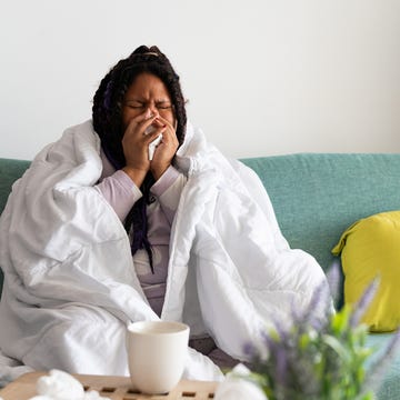 Sick young woman blowing nose while sitting on sofa at home common pneumonia symptoms - young woman sitting on the sofa at home, covered with a blanket, blowing her nose, suffering from a cold or flu