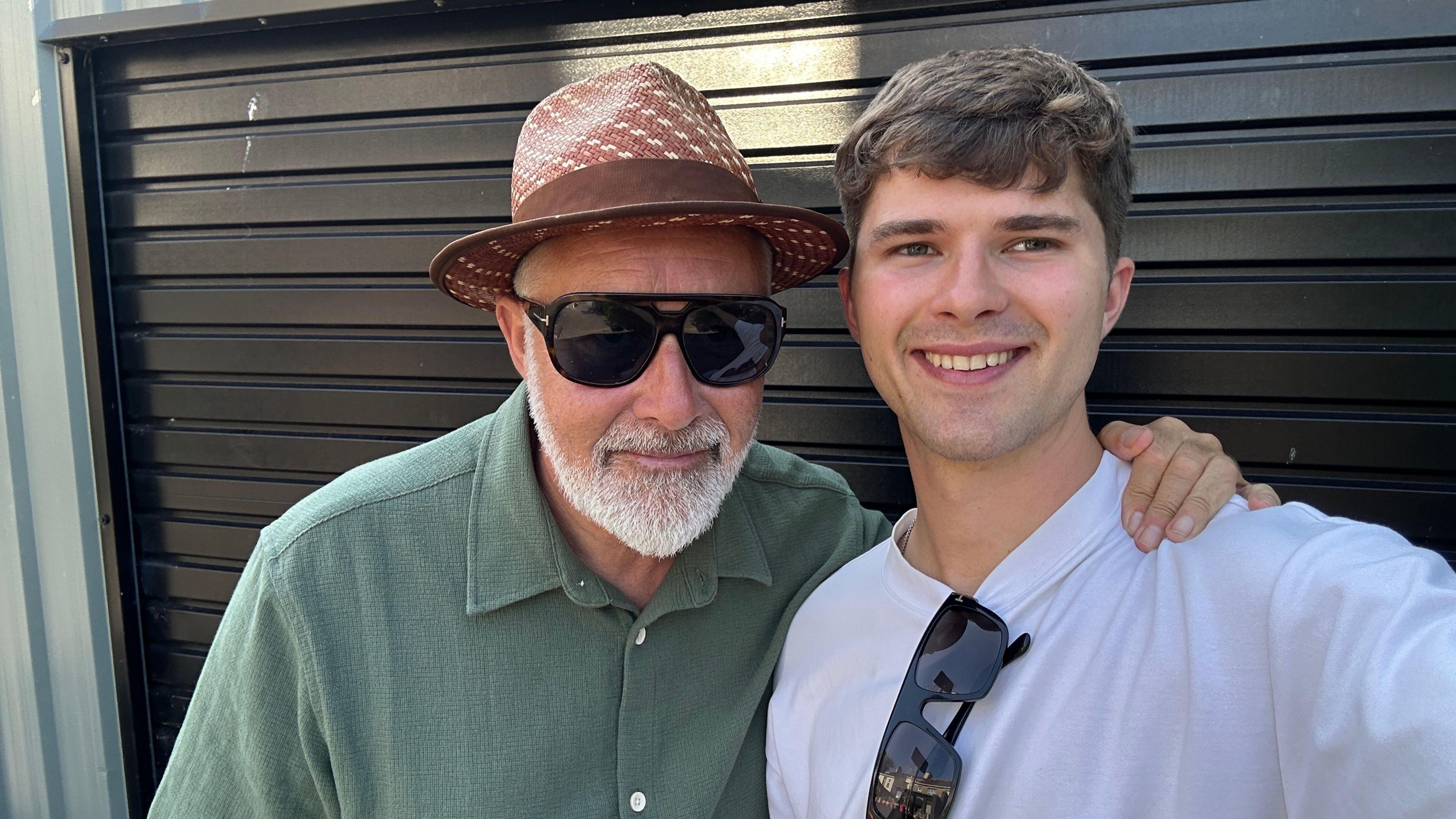 Selfie of Hal Bradley along side his father Terry Bradley. Hal Bradley is to the right, brown hair, white top with sunglasses hanging from it. Terry Bradley is wearing sunglasses and a brown hat, He has a white beard and a green top.