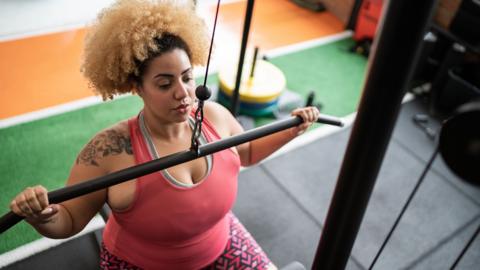 Strock photo shows a person with a flower tattoo on their right shoulder wearing red and pink sportswear pulling down a lateral weights bar in a gym setting.