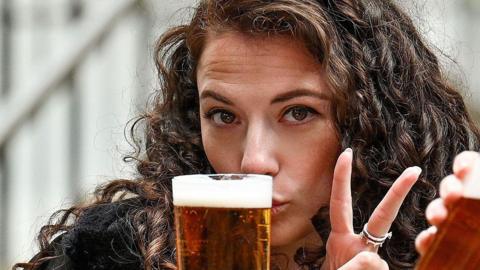A woman and a man sitting outdoors holding pints of Lager. She is making a peace sign and he is drinking, in a casual seating area with steps and railings in the background