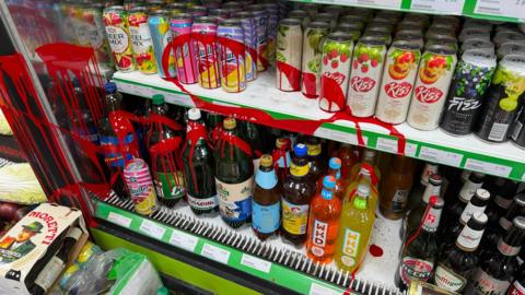 A fridge in a shop, with red paint splattered on items. It is across bottles, and cans.