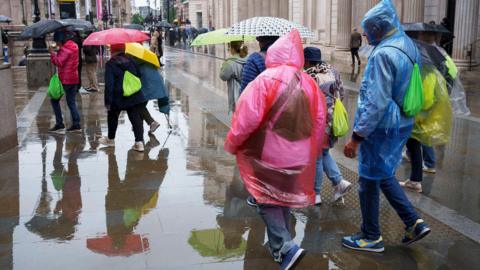 A crowd of people in colourful rain ponchos walk through a rain-soaked street in London. Some are holding umbrellas.