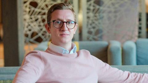 A man wearing glasses and a light pink jumper while seated on a grey sofa in the foreground. One arm rests along the back of the sofa and the other on the armrest. Additional sofas and a patterned room divider are visible in the background of the indoor lounge area.