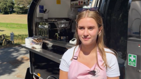 Hariette standing in front of her coffee van. She is wearing a white T-shirt and a pink apron.