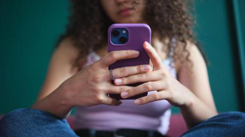 A young woman, whose face cannot be seen, sits cross-legged while holding and using her smartphone. She is wearing blue jeans and a lilac vest top.