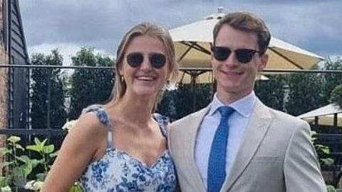 Alexander Cromie and Marie Ebert stand arm in arm and smile. Alexander is wearing a tan suit and sunglasses. Marie is also wearing sunglasses and a floral dress.