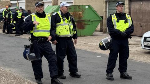 Three police officers are pictured standing in a road. They are wearing yellow vests and all have caps on
