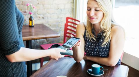 Woman sitting in a cafe with a coffee cup on the table in front of her puts her phone on a payment terminal held by a waitress.
