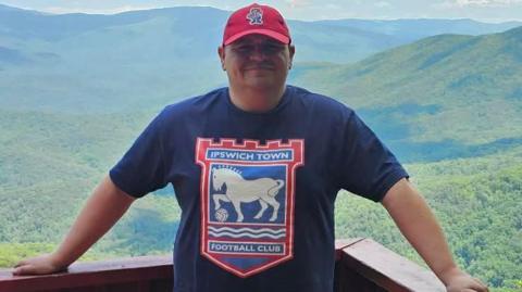 A man stands on a balcony in front of a large expanse of trees and hills. He wears a navy T-shirt with the Ipswich Town logo on it as well as a red cap.