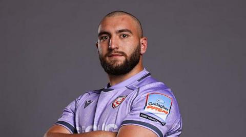 Archie McArthur standing looking at the camera with his arms folded. He is wearing purple Gloucester kit