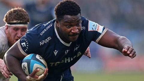 Sale Sharks hooker Nathan Jibulu, in dark blue Sale shirt with white trim, carries the ball as Bath's Harvey Cuckson, in white and with a black-and-white headband, reaches out to grab him from behind