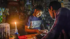 একটি দোকানে মোমবাতি জ্বালিয়ে কাজ করছেন একজন, সামনে আরেকজন দাঁড়িয়ে আছেন