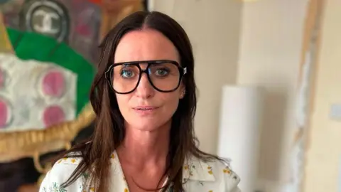 A woman with long dark hair looks at the camera. She is wearing large black glasses